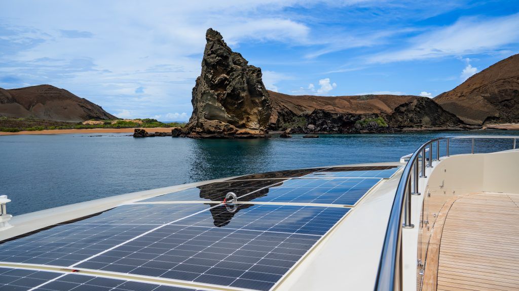 Solar panels on a yacht deck overlooking calm blue water and a jagged rocky island under a blue sky.